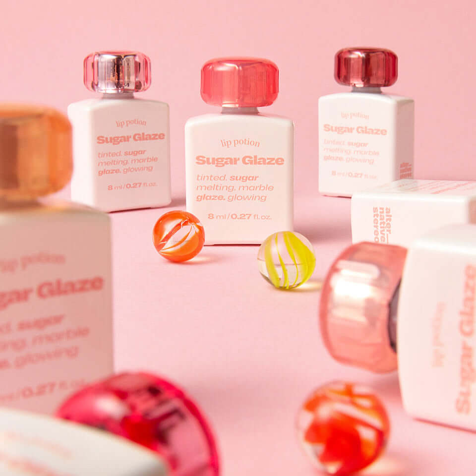 Display of ALTERNATIVE STEREO Lip Potion Sugar Glaze bottles with colorful design on a pink background, highlighting product aesthetics.