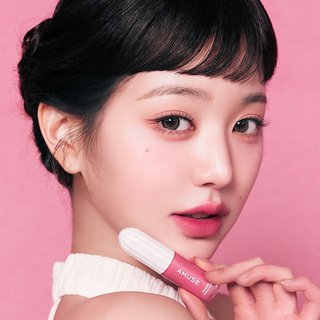 Model wearing AMUSE Powder Velvet Tint in pink shade, holding the product in front of a matching pink background for a vibrant look.