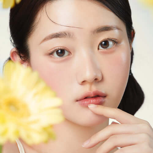 Model showcasing clear skin after using APRILSKIN Calendula 10% PHA+AHA Resurfacing Serum with yellow flowers in foreground.
