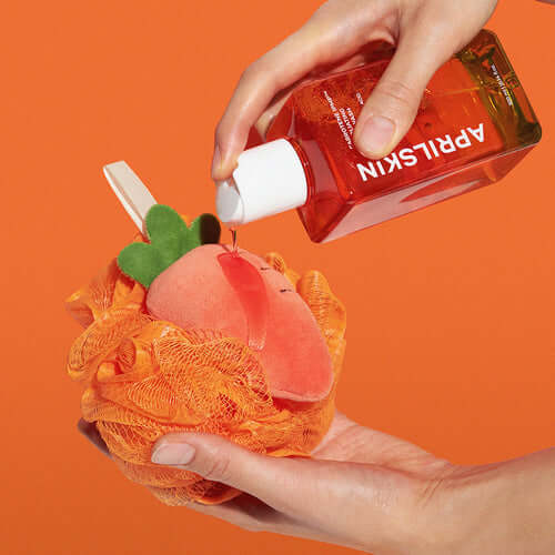 Hands using APRILSKIN Carrotene IPMP Exfoliating Body Wash with orange loofah on vibrant orange background