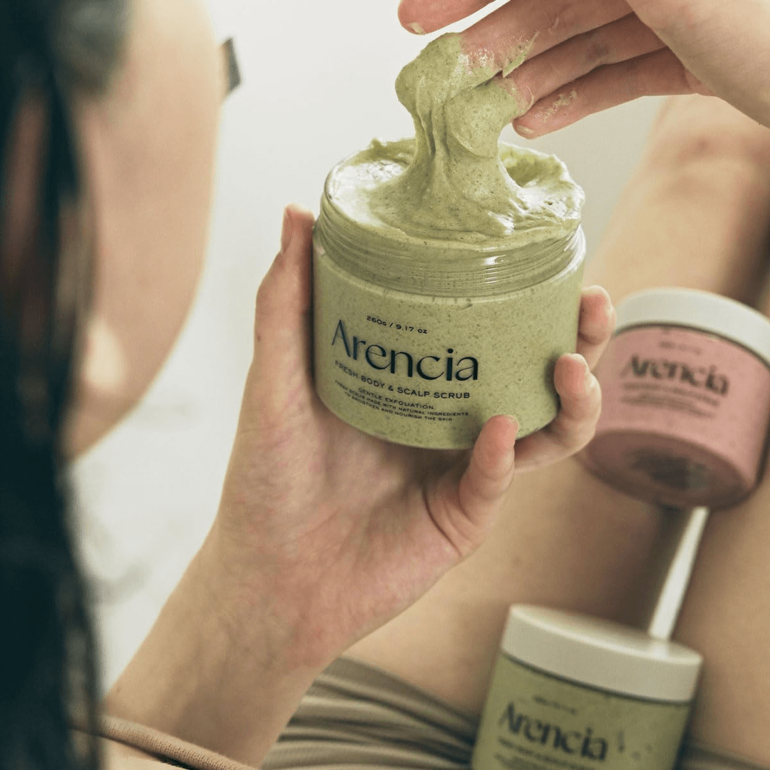 Person using ARENCIA Fresh Cloud Body Scrub 260g, scooping creamy texture from jar, with pink and green containers in background.