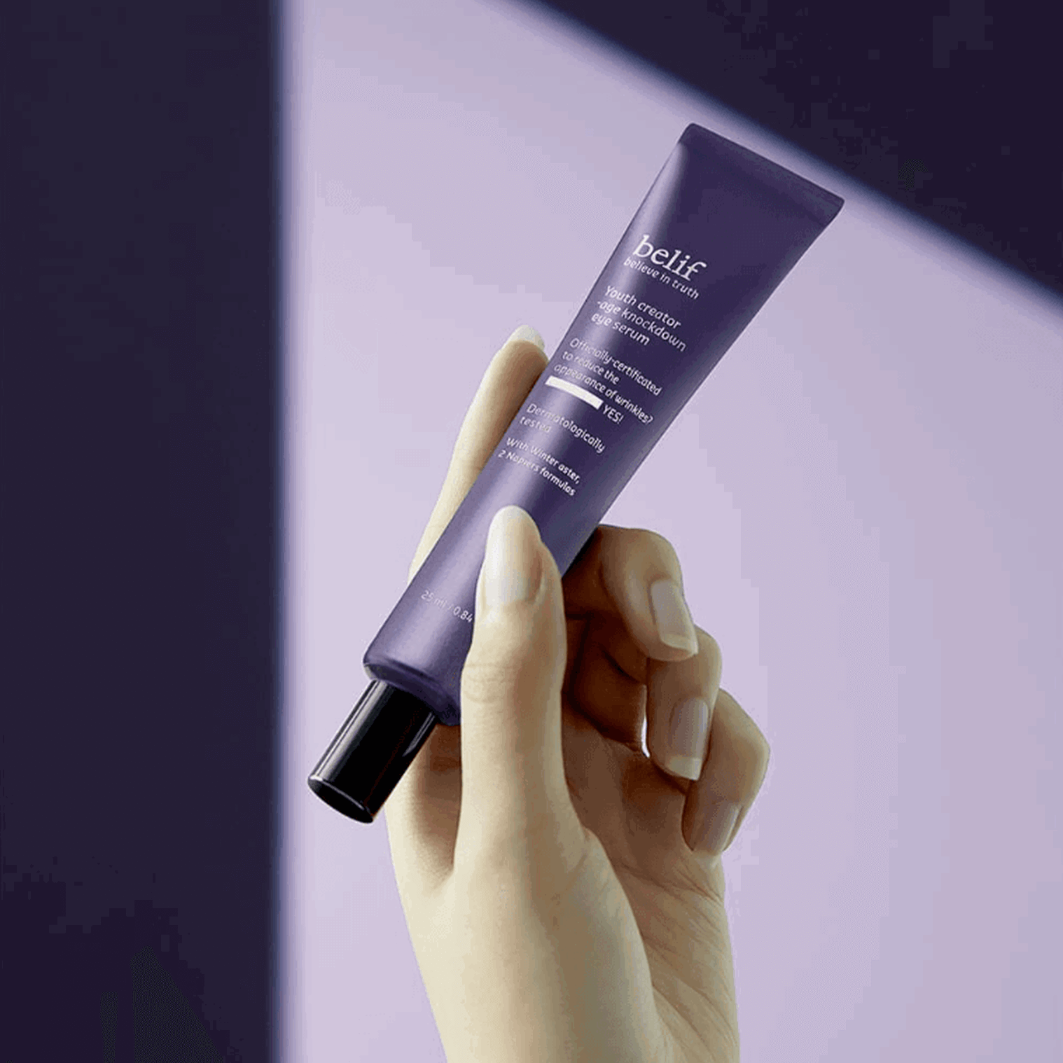 Hand holding BELIF Age Knockdown V Eye Serum 25ml against a purple background.