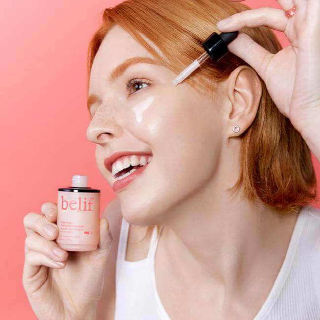 Smiling woman applying BELIF Super Drops Peptide Firming Serum 30ml with dropper on pink background