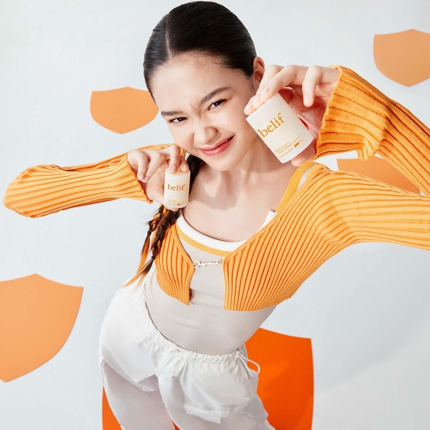 Person holding BELIF Super Knight Multi Vitamin Lipcerin products wearing orange sweater against orange and white background.