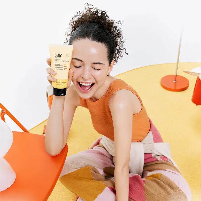 Woman holding BELIF Super Knights bright Toning Vegan Pack Cleanser 150ml, smiling on a colorful background