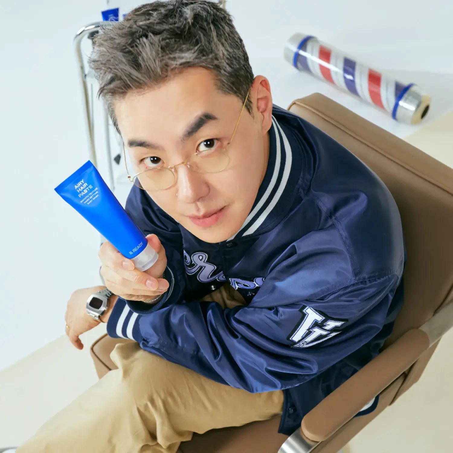 Man holding B.READY Airy Hair Paste 80g in a blue tube, sitting on a chair with a barbershop pole in the background.