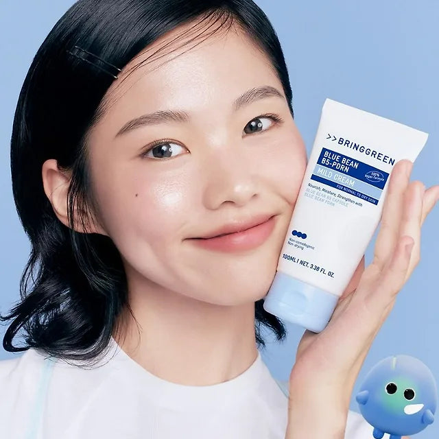 Smiling woman holding BRINGGREEN Blue Bean B5-PDRN Mild Cream 100ml with mascot, showcasing the product's packaging.