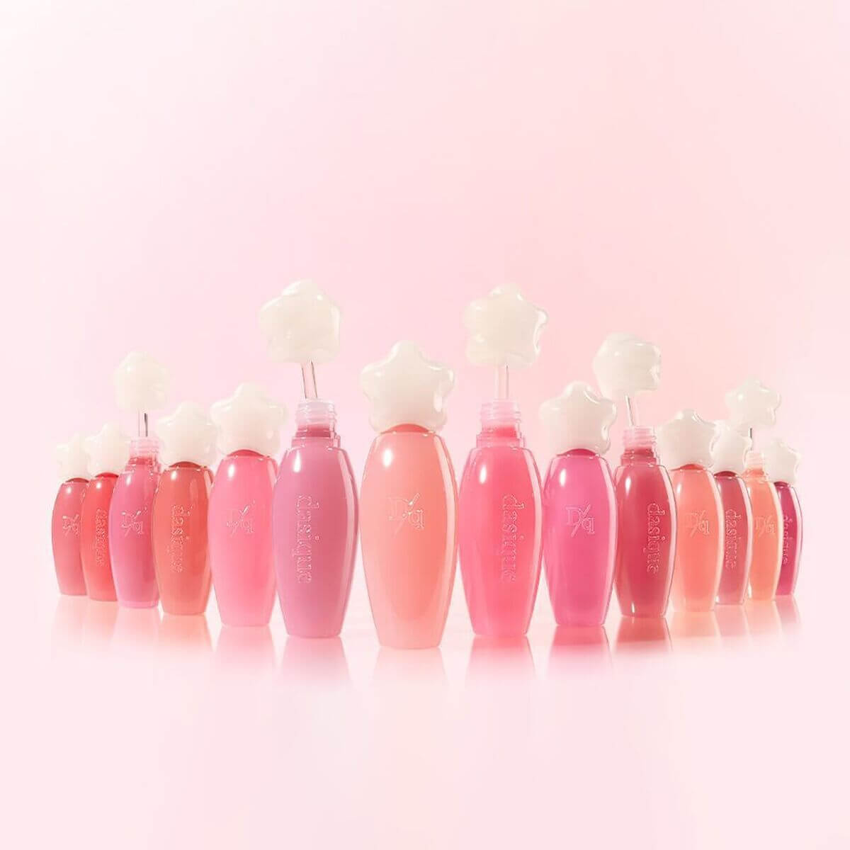 Lineup of DASIQUE Pure Water Lip Gloss bottles in various shades, set against a pink background. Stylish packaging with flower-shaped caps.