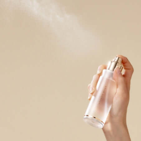 Hand holding DEAR DAHLIA Endless SKin Setting Spray bottle, spraying mist against a beige background.
