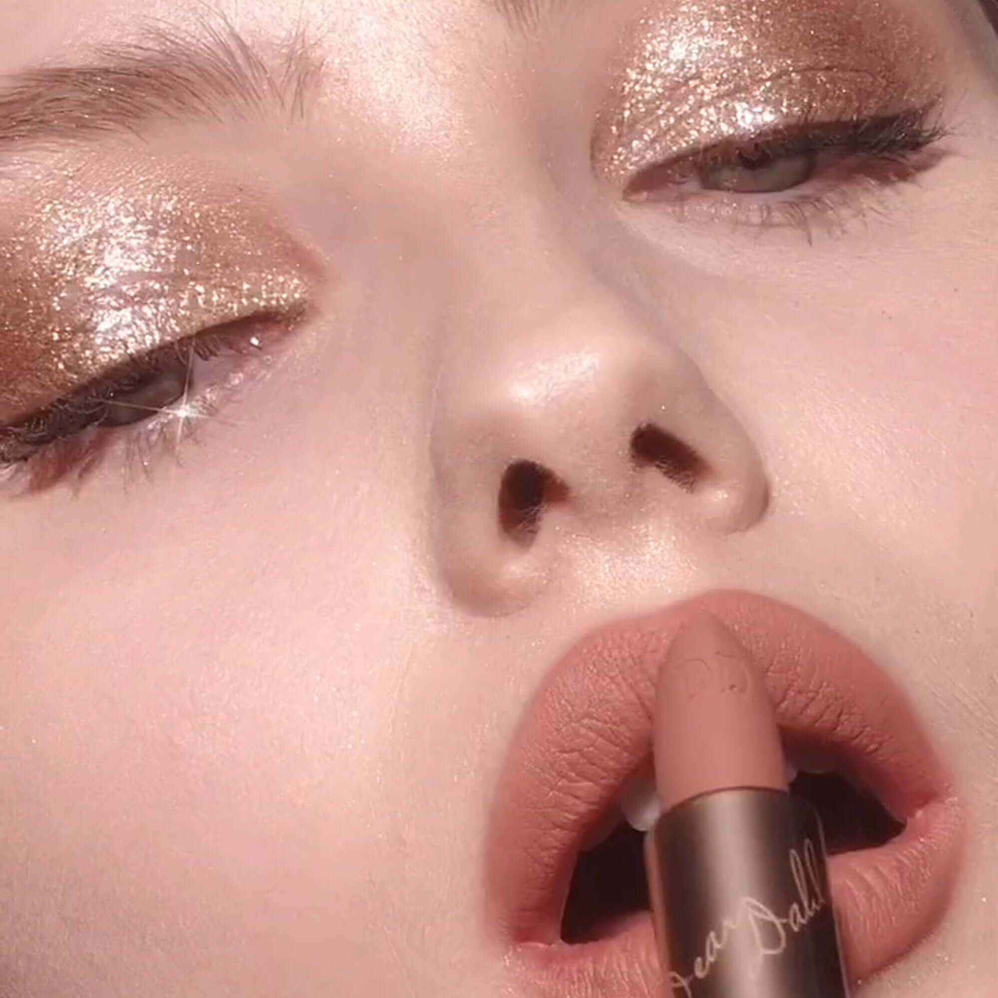Model applying DEAR DAHLIA Lip Paradise Effortless Matte Lipstick in nude shade with shimmering eyeshadow makeup.