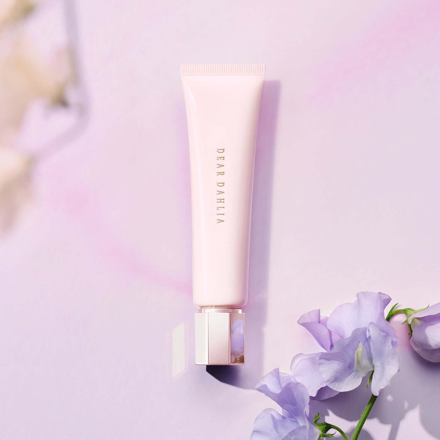DEAR DAHLIA Prime Layer Blurring Primer tube on a pastel pink background with purple flowers for beauty and makeup enthusiasts.