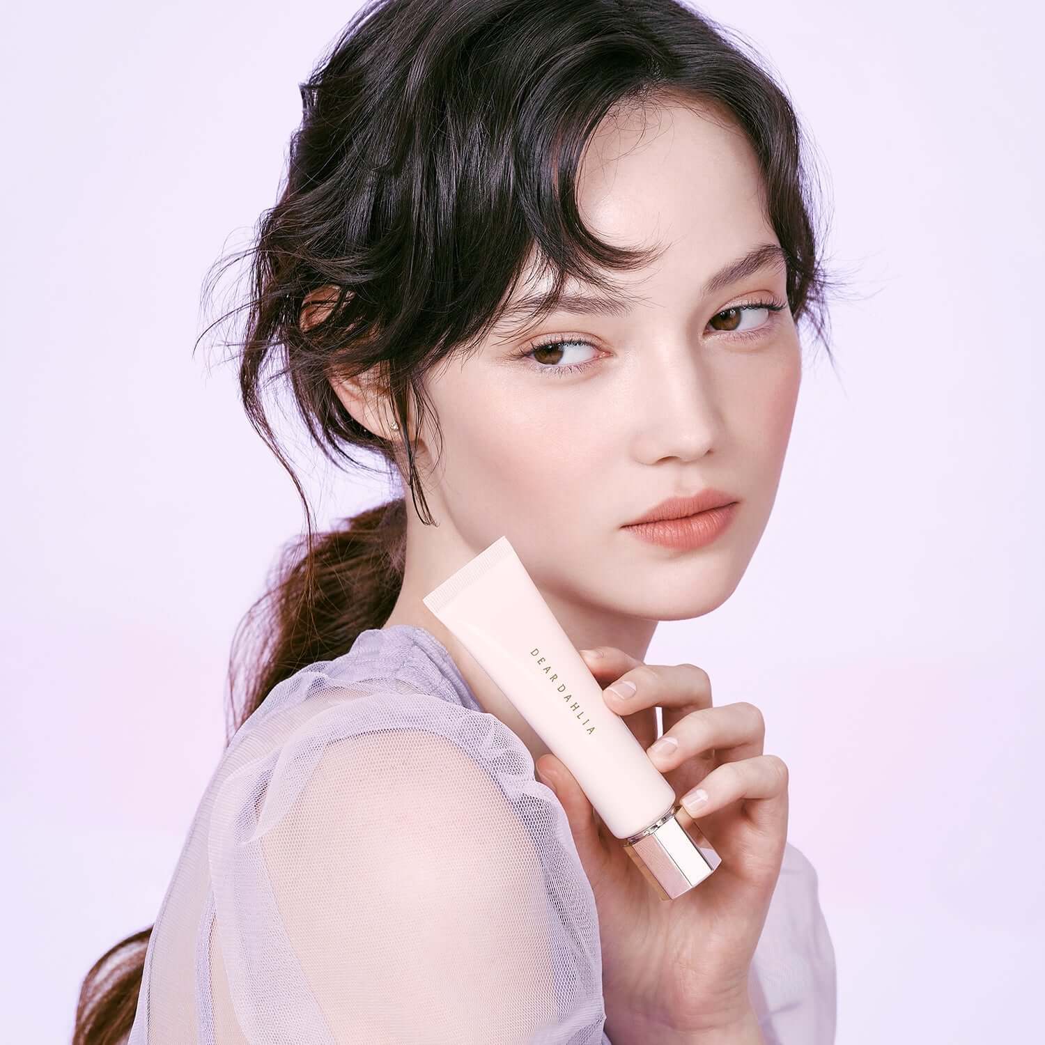 Model holding DEAR DAHLIA Prime Layer Blurring Primer, enhancing soft-focus effect for flawless skin, perfect for beauty enthusiasts.