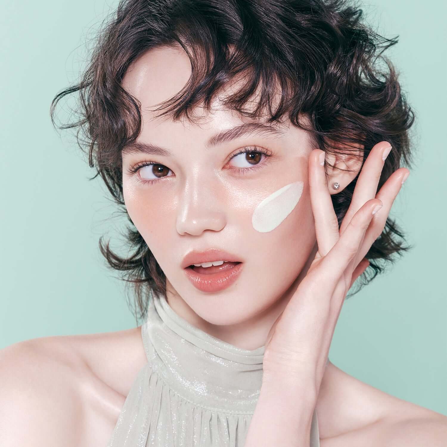 Woman applying DEAR DAHLIA Prime Layer Correcting Primer to her face against a soft green background.