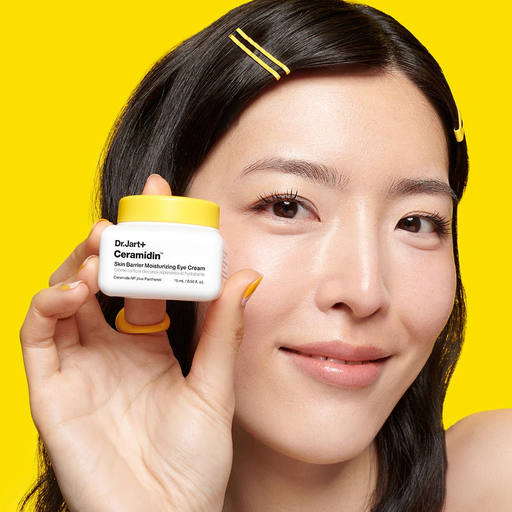 DR.JART+ Ceramidin Skin Barrier Moisturizing Eye Cream 15ml bottle held by model against yellow background.