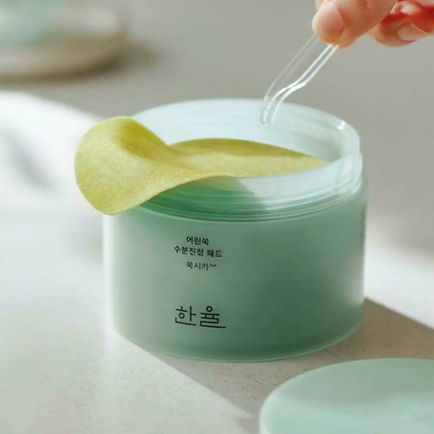 Close-up of HANYUL Young Mugwort Moisturizing Soohting Pad container with pads being picked up by tweezers, showing product texture.