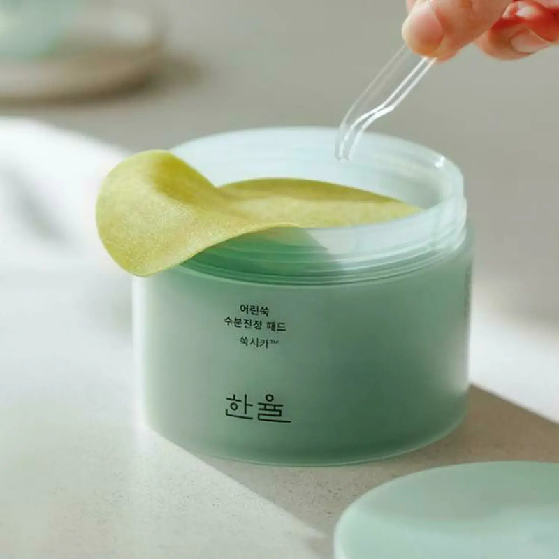 Close-up of HANYUL Young Mugwort Moisturizing Soohting Pad container with pads being picked up by tweezers, showing product texture.