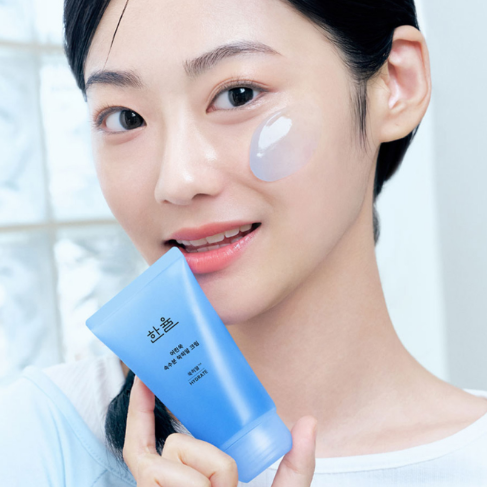 Smiling person holding HANYUL Young Artemisia Extract Moisturizing Cream 60ml, applying cream on cheek.