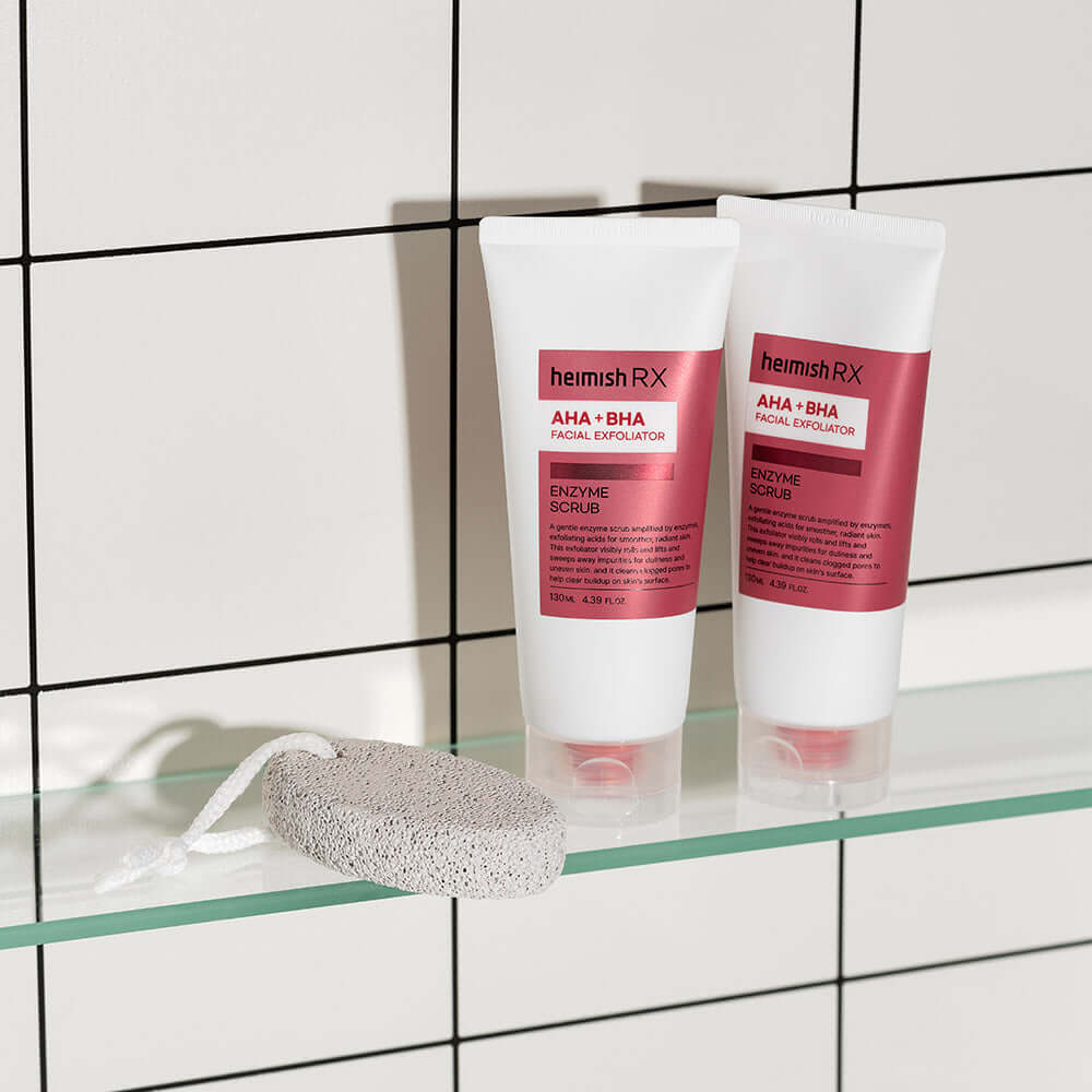HEIMISH RX AHA BHA Enzyme Scrub 130ml displayed on a glass shelf with a pumice stone in a tiled bathroom setting