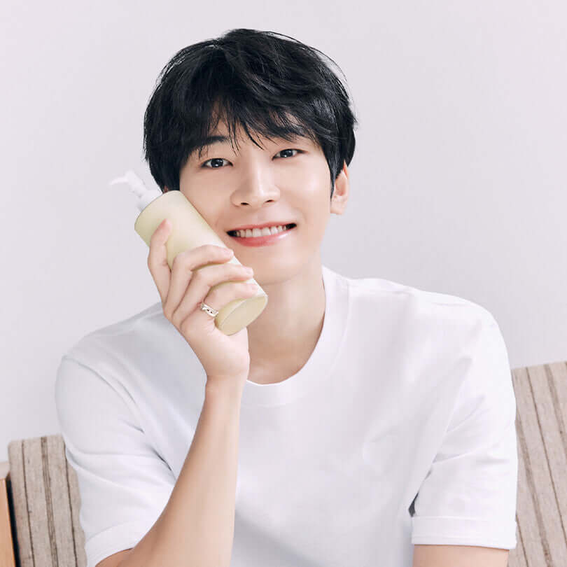 Person holding a bottle of HUXLEY Body Lotion Blue Madina Tangerine, smiling in a white t-shirt.