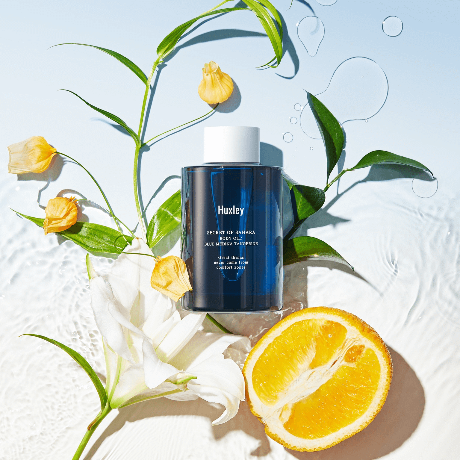 HUXLEY Body Oil Blue Medina Tangrine with fresh tangerine slices and floral accents against a translucent backdrop