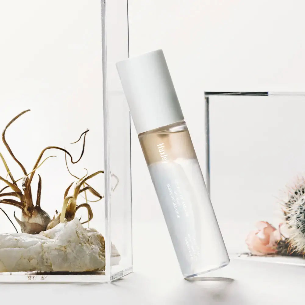 Bottle of HUXLEY Oil Mist Sense Of Balance next to decorative plants on a white background.