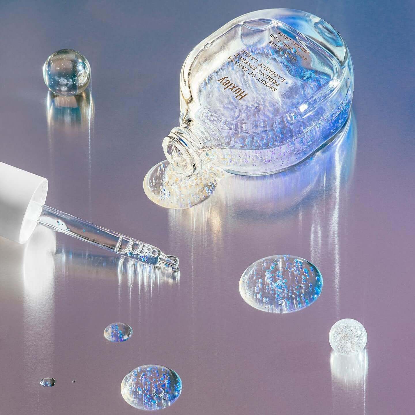 HUXLEY Priming Essence Rediance Layer bottle and dropper with iridescent liquid on metallic surface, highlighting radiant skincare.
