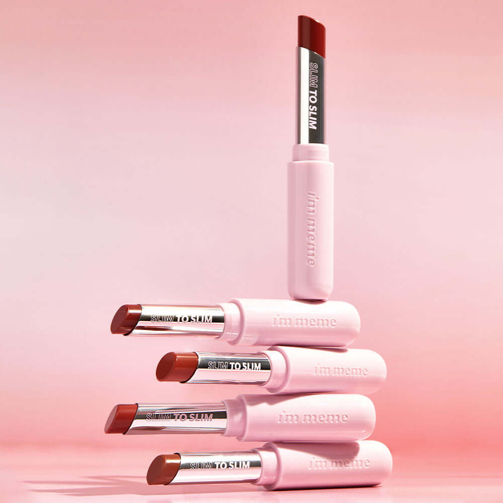 Stack of IM MEME Slim to Slim Hyaluronic Gloss Fit Lipsticks displayed on a pink background.