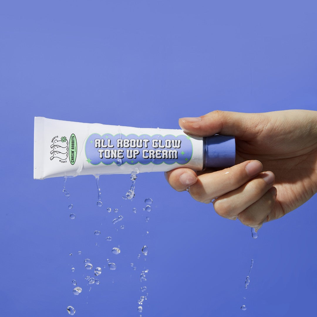 Hand holding IT'S SKIN All About Glow Tone Up Cream against a blue background, with water droplets cascading around the product.