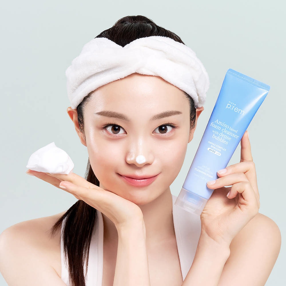 Person holding MAKE P:REM Safe Me Amino Refresh Cleansing Foam 150ml beside face, with cleansing foam on nose and headband on.
