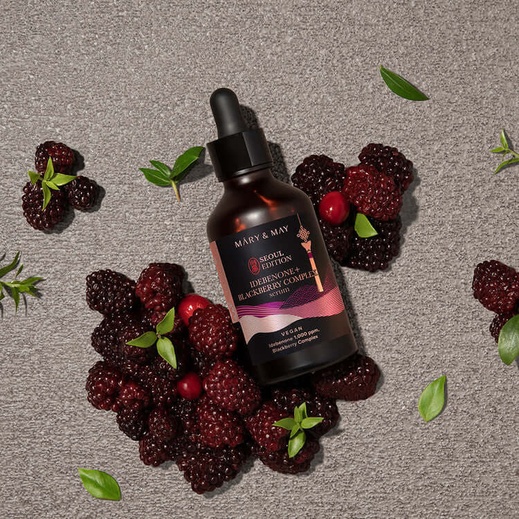 MARY & MAY Idebenone+Blackberry Complex Serum bottle surrounded by fresh blackberries on a textured surface