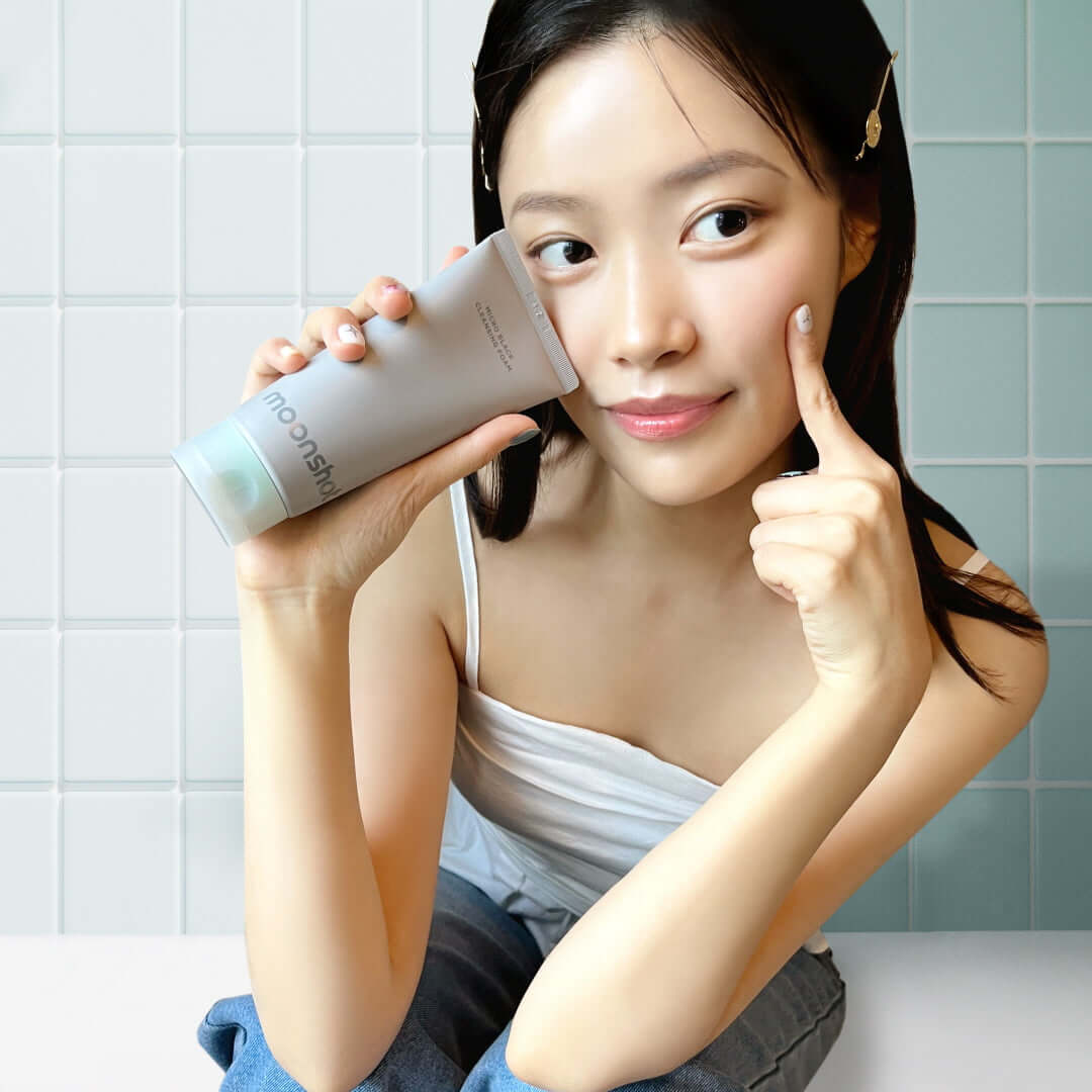 Woman holding MOONSHOT Micro Black Cleansing Foam tube against her cheek in a bathroom.