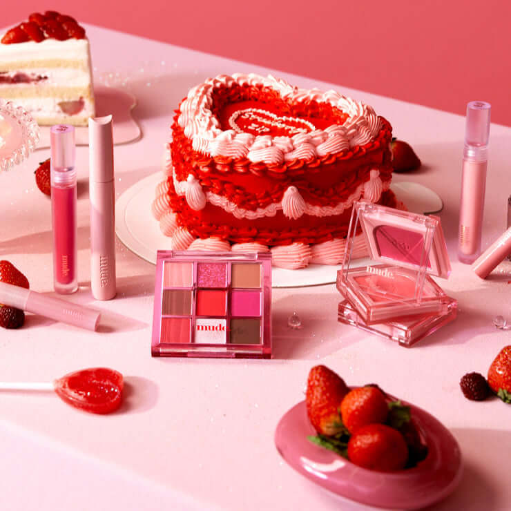 MUDE Feel Lively Eyeshadow Palette with pink and red shades surrounded by desserts and makeup items on a pink table.
