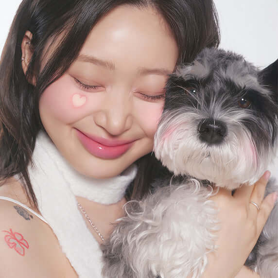 Smiling woman with heart decorations on cheeks hugging a fluffy dog showing PERIPERA Syruppy Talk Cheek effect.
