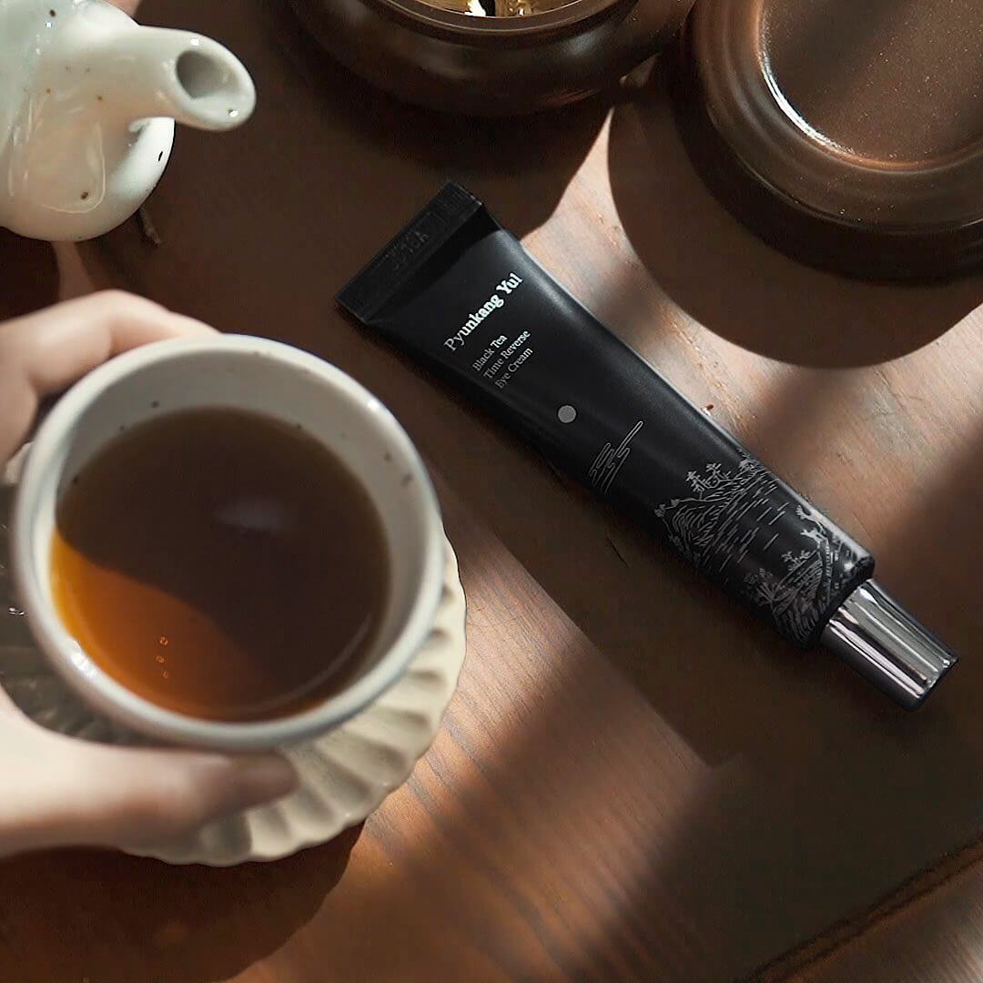 PYUNKANG YUL Black Tea Time Reverse Eye Cream 25ml placed next to a cup of tea on a wooden surface