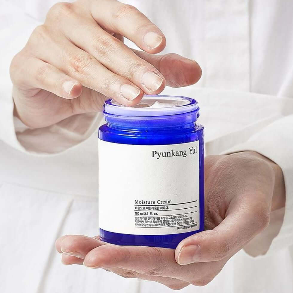Hands holding an open jar of PYUNKANG YUL Moisture Cream 100ml with a blue container.