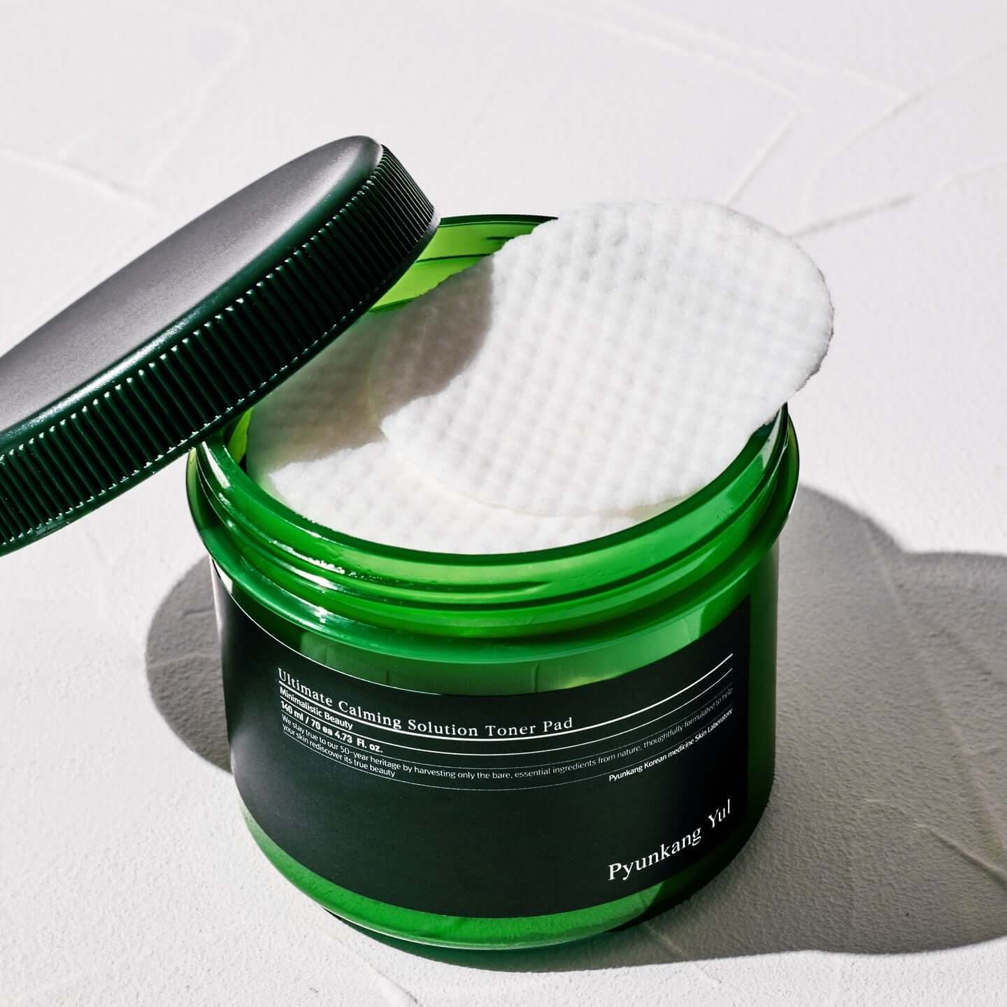 Open jar of PYUNKANG YUL Ultimate Soothing Solution Toner Pads 70 sheets 140ml with white pad halfway out on white background