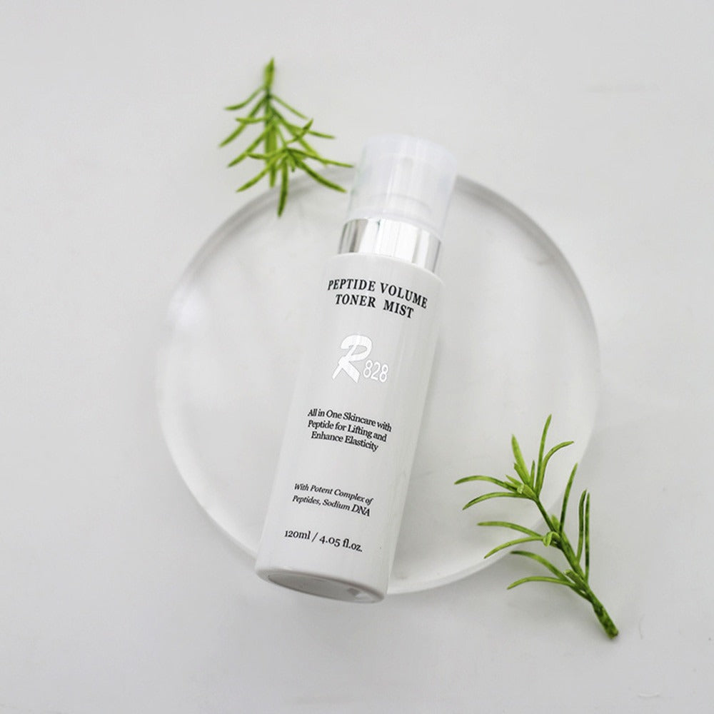 R828 Peptide Volume Toner Mist on a clear display with green leaves, 120ml bottle for skin care hydration and peptide infusion.