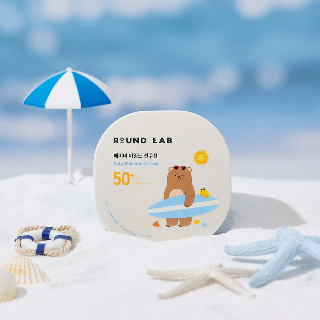 ROUND LAB Baby Mild Sun Cushion 16g with SPF 50 packaging displayed on sandy beach with small umbrella and beach toys.
