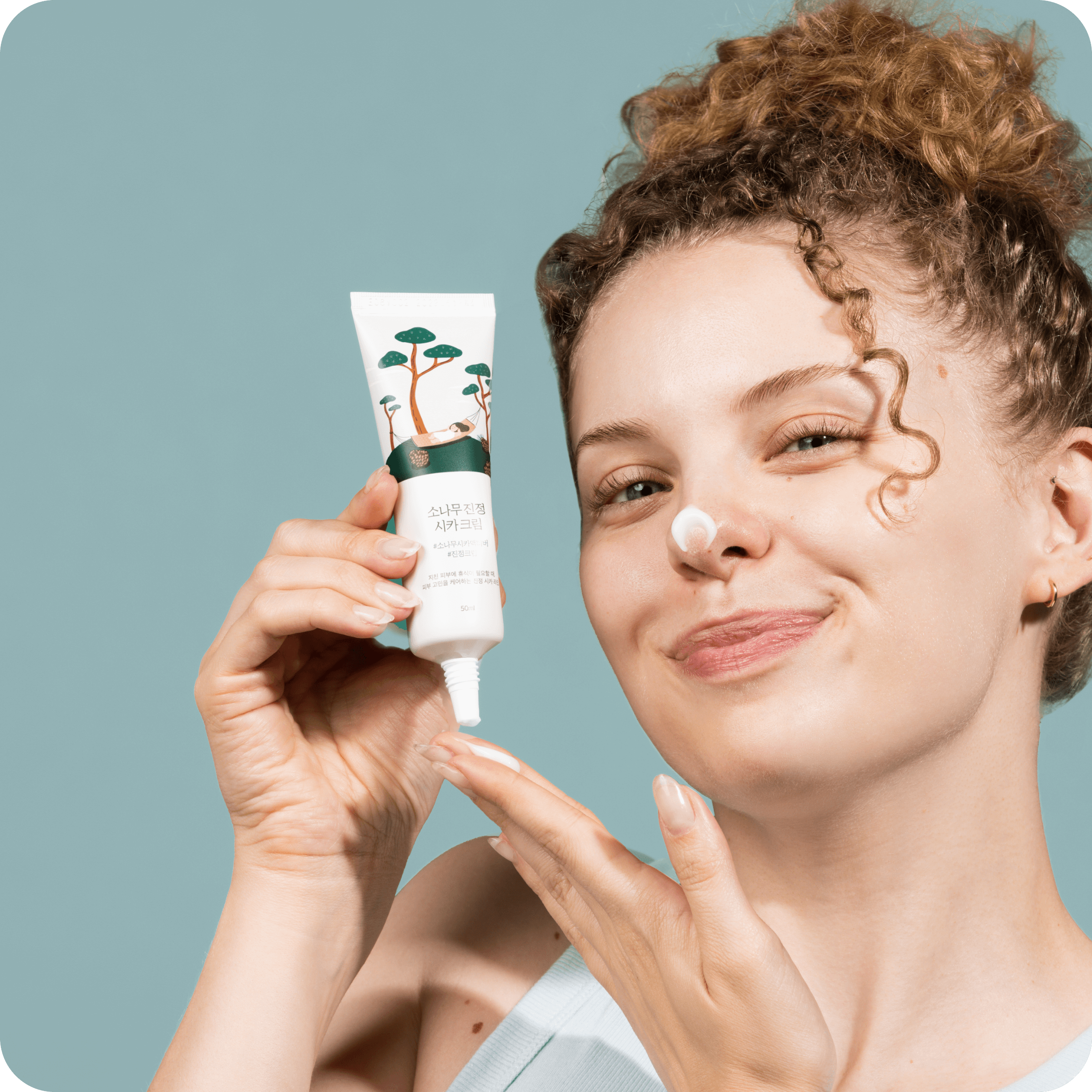 Woman applying ROUND LAB Pine Calming Cica Cream 50ml to her face with a smile, showcasing the skincare product's calming effects.