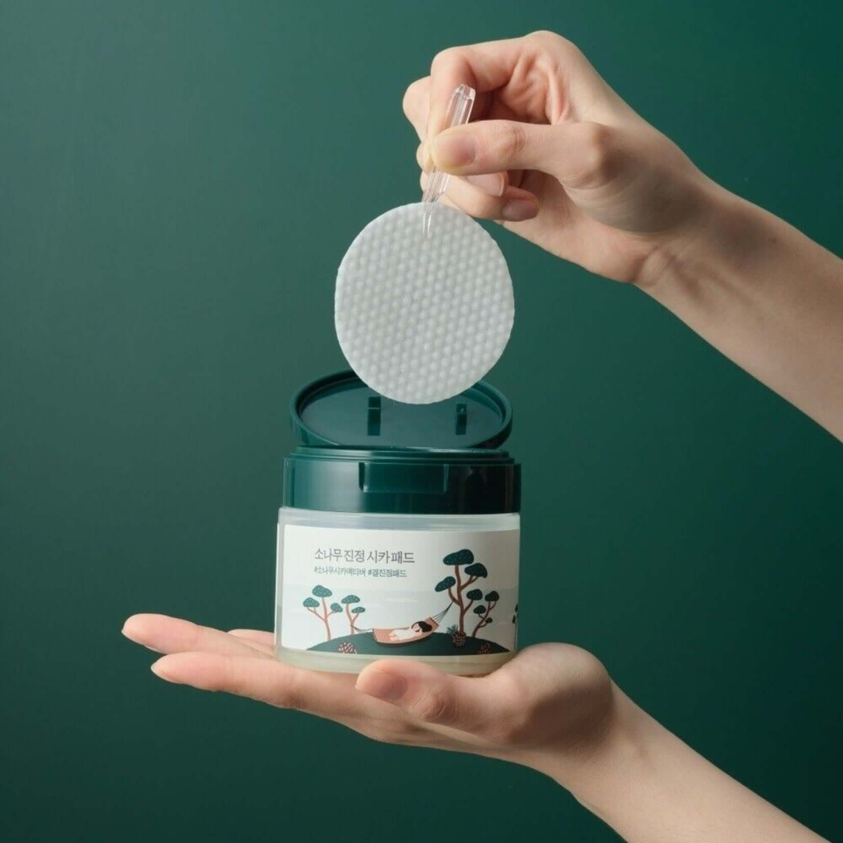 Hands holding ROUND LAB Pine Calming Cica Pads 50 sheets with open jar, showcasing textured pad against green background
