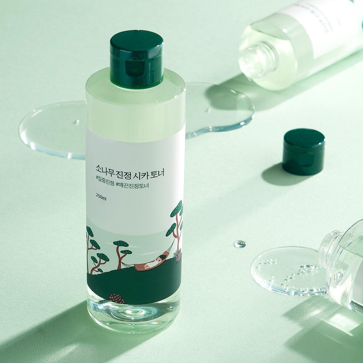 ROUND LAB Pine Calming Cica Toner 250ml bottle on a light green background with scattered toner and open caps. Skincare product image.