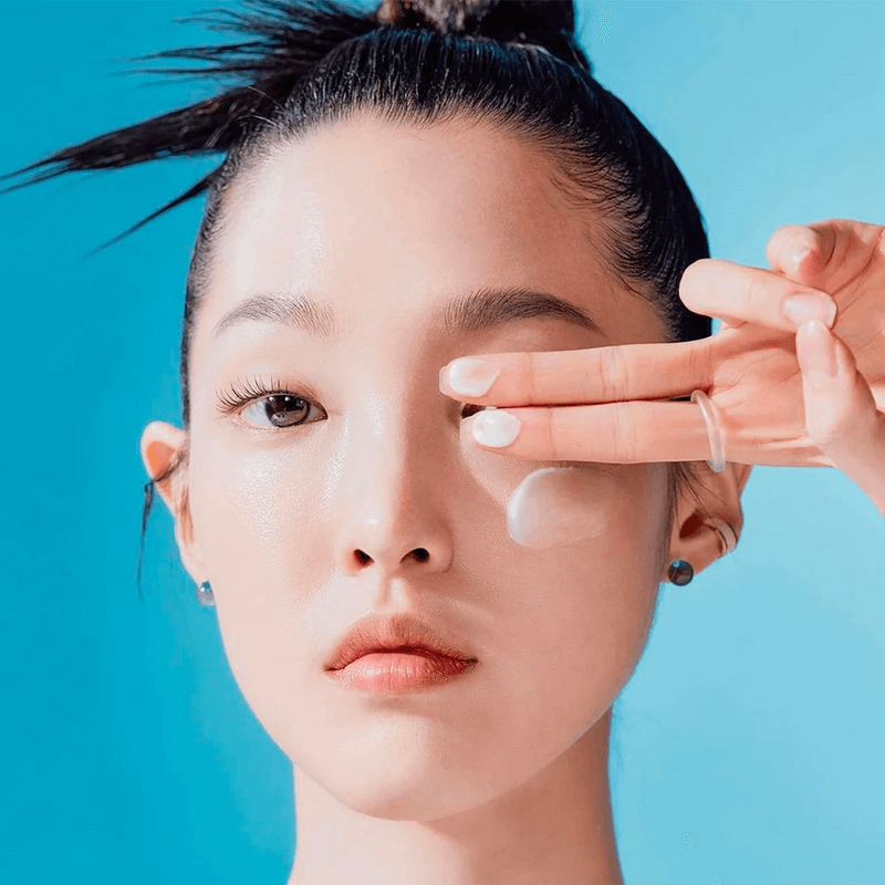 Woman applying TOCOBO Sun Care Mini Duo sunscreen to face, close-up with blue background