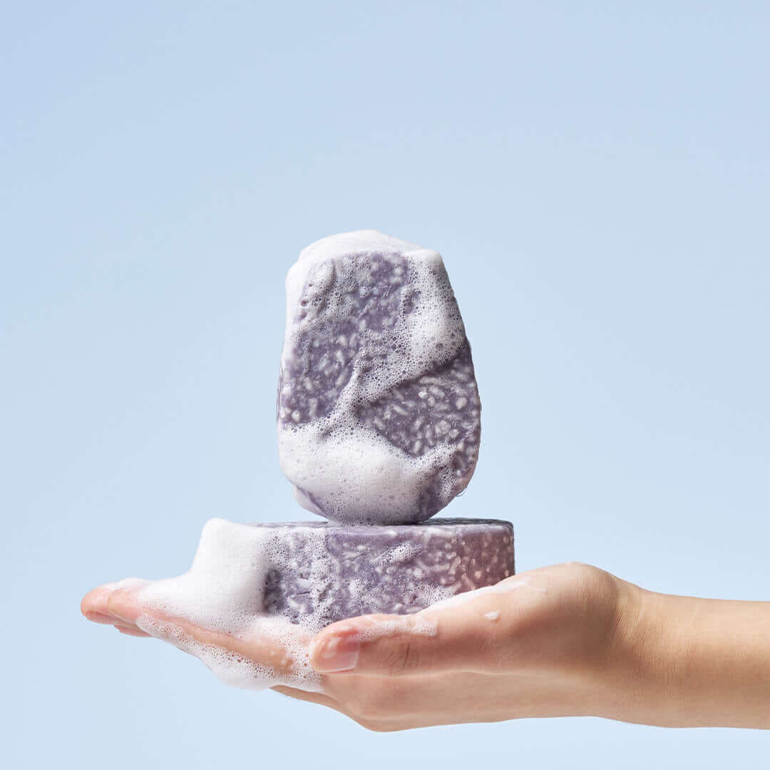 Hand holding two lathered URANG Daily Oil Free Shampoo Bars against a blue background.