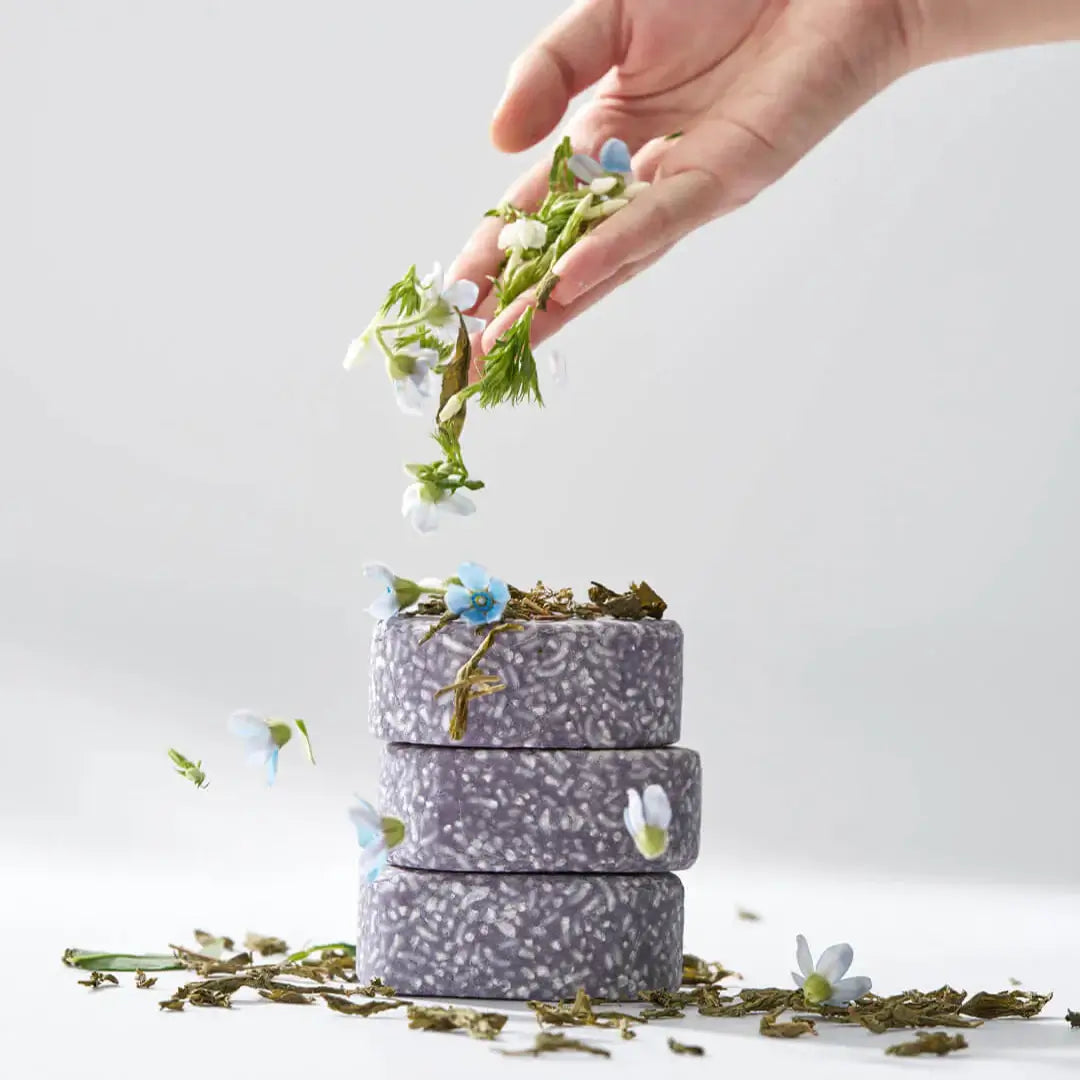 A hand decorating stack of URANG Daily Oil Free Shampoo Bar with flowers and leaves in a minimalist setting.