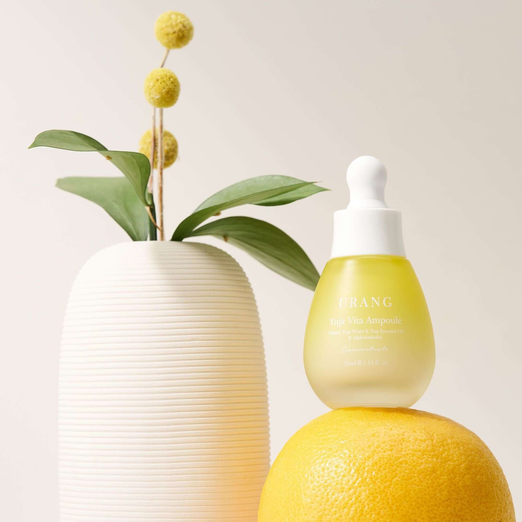 URANG Yuja Vita Ampoule bottle placed on a citrus fruit next to a modern vase with yellow flowers and green leaves.
