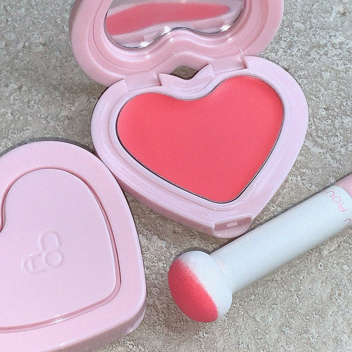 AOU Soft Cream Blush in heart-shaped compact with applicator, showcasing its vibrant pink hue on a textured background.
