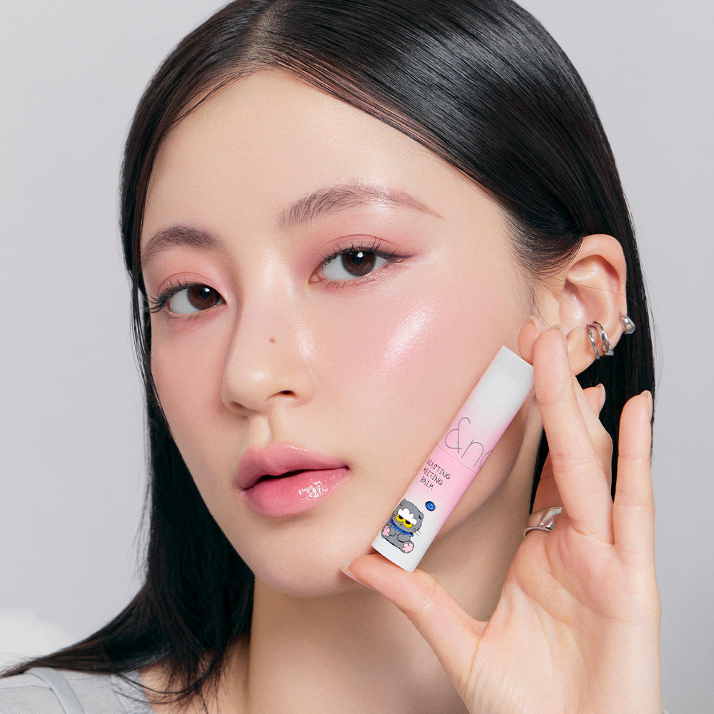 Woman holding ROMAND x Joanne Friends Glasting Melting Balm with pink packaging and cartoon character design.