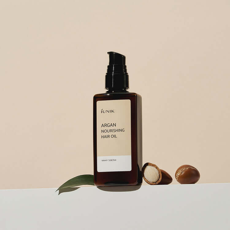 Bottle of iUNIK Argan Nourishing Hair Oil 100ml with argan nuts and a leaf on a neutral background