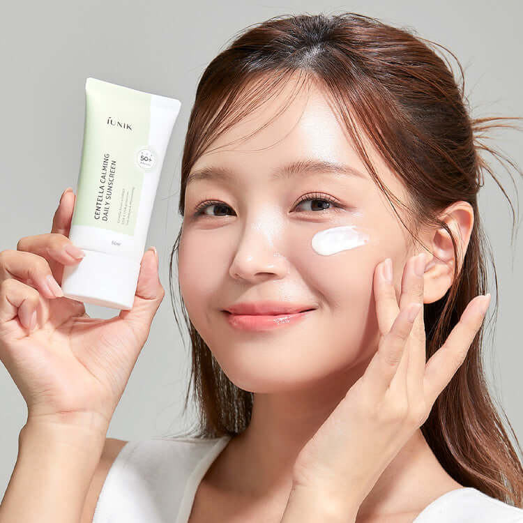 Smiling woman applying iUNIK Centella Calming Daily Sunscreen 60ml to her face, holding the product with skincare benefits.