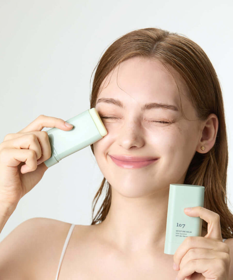 Woman applying 107 Moisture Balm Mild Sun Stick SPF50+PA++++ to her face, showcasing skincare routine with sunscreen protection.
